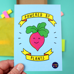 A hand holds up an A6 small art print. The background is blue and in the centre is an ilustration of a pink root vegetable with a cute smiling face, with green leaves for hair. Around this illustration are yellow banners containing the words 'Powered by Plants'