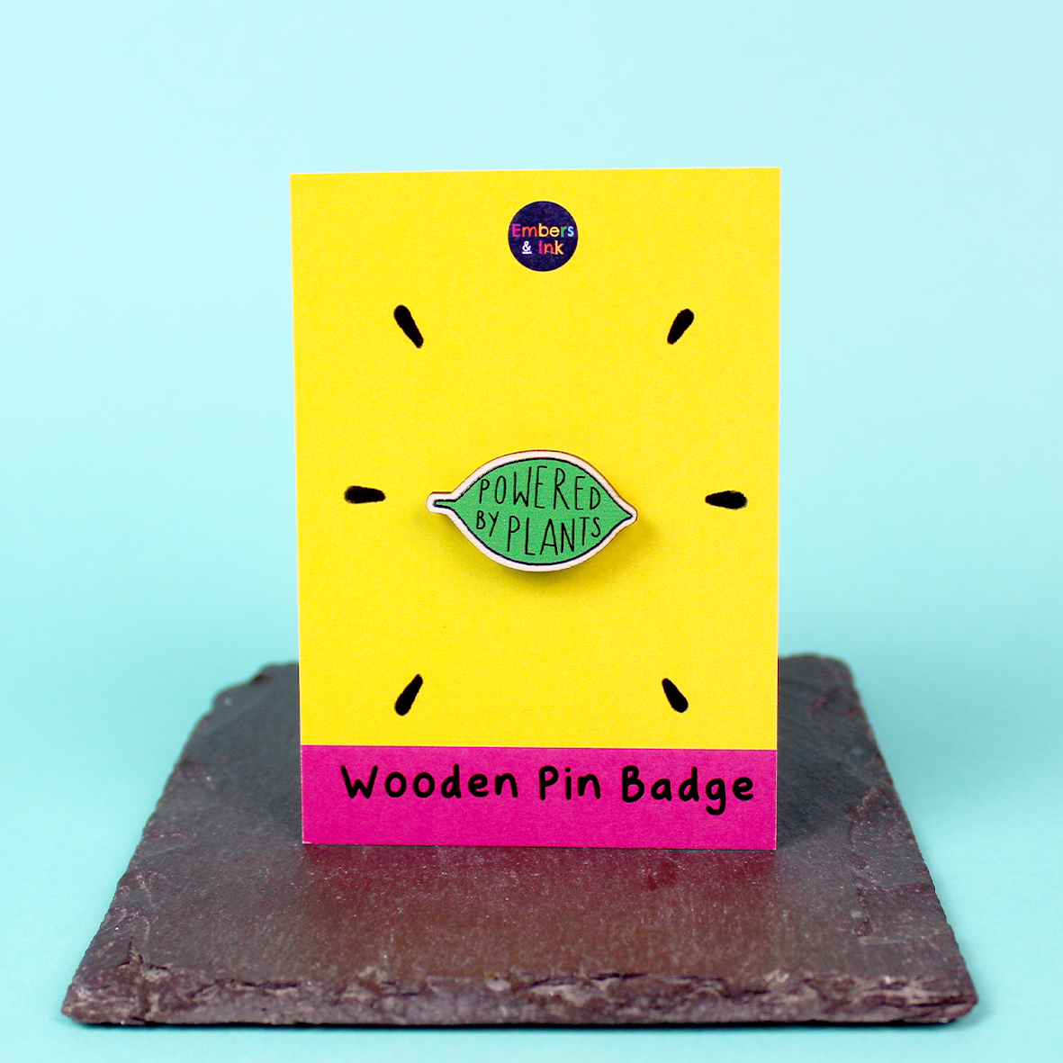 A wooden pin badge in the shape of a green leaf contains the words Powered by Plants. The badge is on a yellow and pink backing board with the words 'wooden pin badge' along the bottom