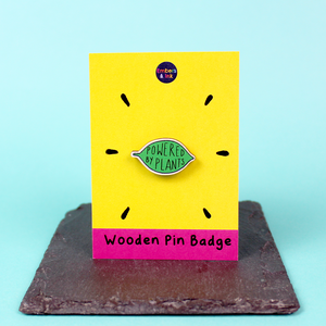 A wooden pin badge in the shape of a green leaf contains the words Powered by Plants. The badge is on a yellow and pink backing board with the words 'wooden pin badge' along the bottom
