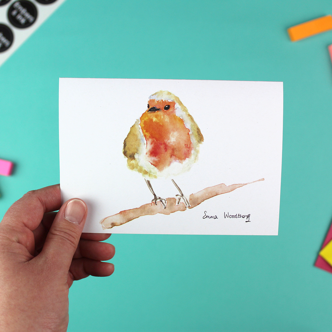 A hand holds a landscape-orientated greetings card featuring a watercolour image of a Robin on a branch.