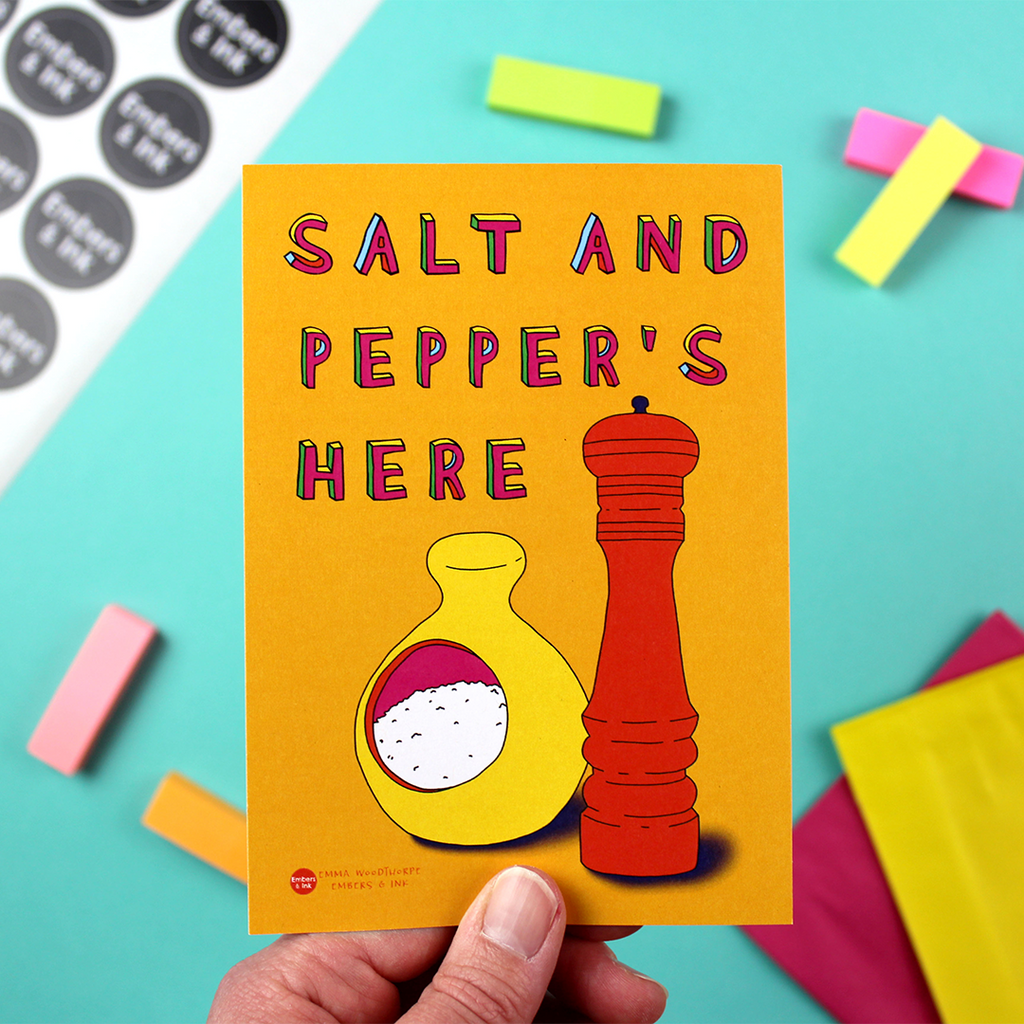 A hand holds up a portrait-orientated small art print of A6 dimensions. It has an orange background and an illustration of a yellow salt pig and an orange pepper mill. above it are the words Salt and Pepper's Here.