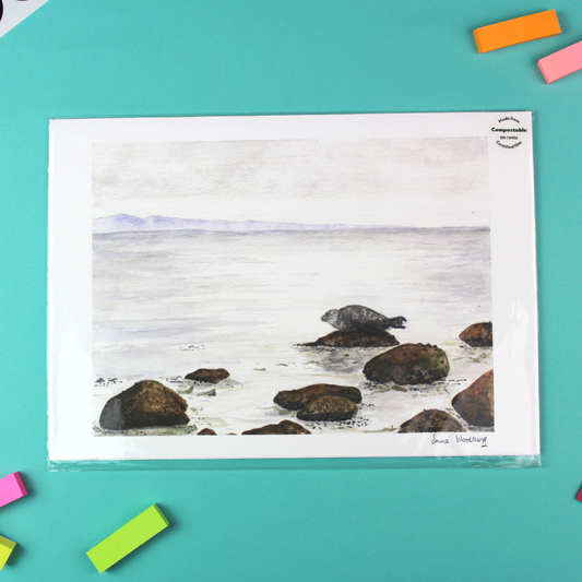 The image shows a landscape orientated print of a watercolour seascale. in the distance across the water are purple mountains, and in the foreground a seal basks on a rock.