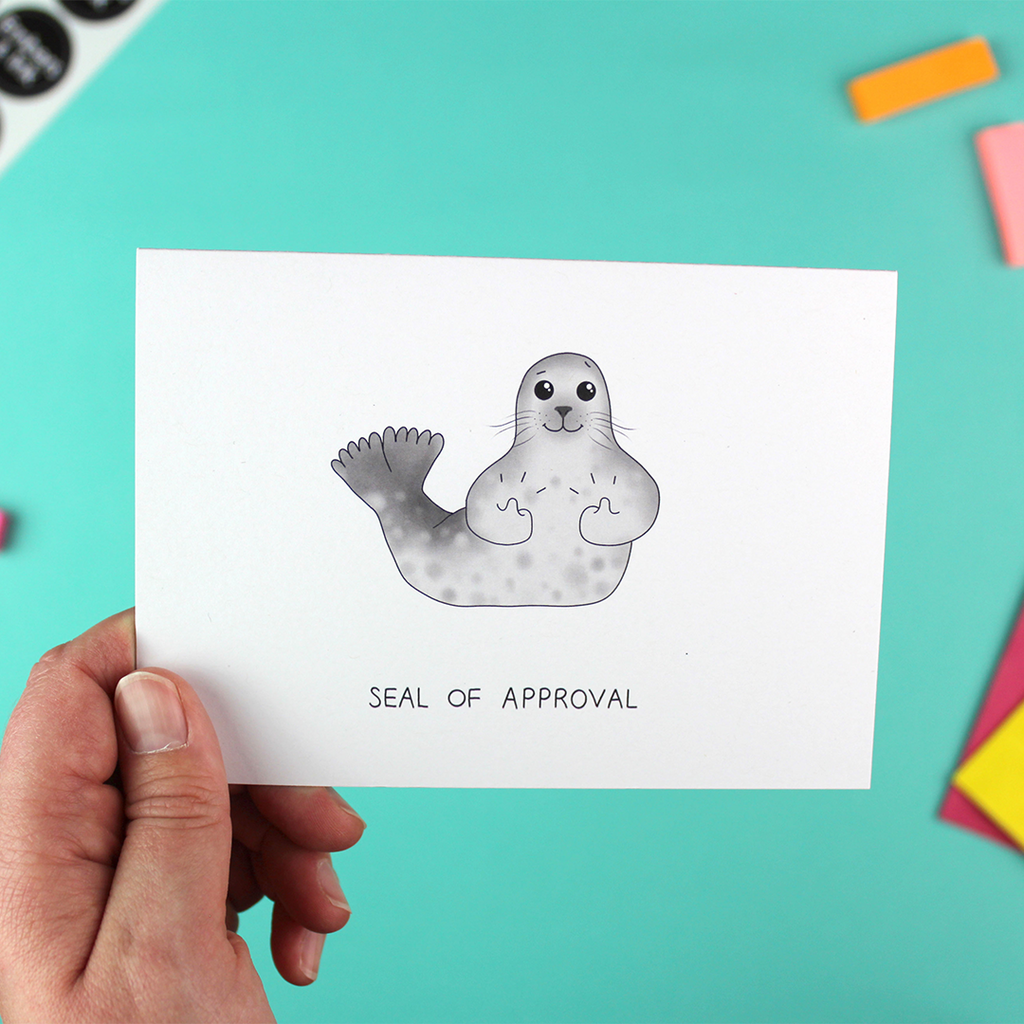A hand holds a landscape orientated greetings card with an illustratio of a seal with its thumbs up. Underneath are the words 'seal of approval'