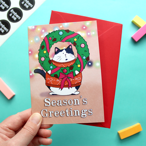 A hand holds a card with an illustration of a cat wearing a Christmas jumper and holding a Christmas wreath