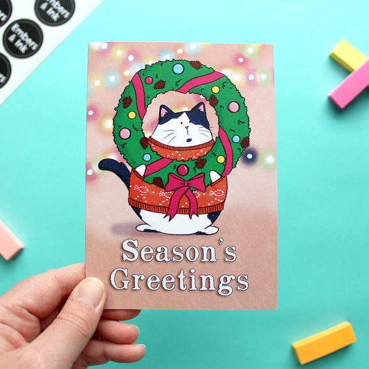 A hand holds a card with an illustration of a cat wearing a Christmas jumper and holding a Christmas wreath