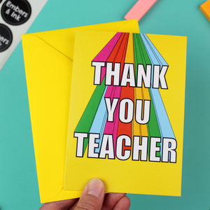 A hand holds a bright and colourful Greetings Card and a yellow envelope. The card has a yellow background and rainbow coloured 3D lettering that reads Thank You Teacher.