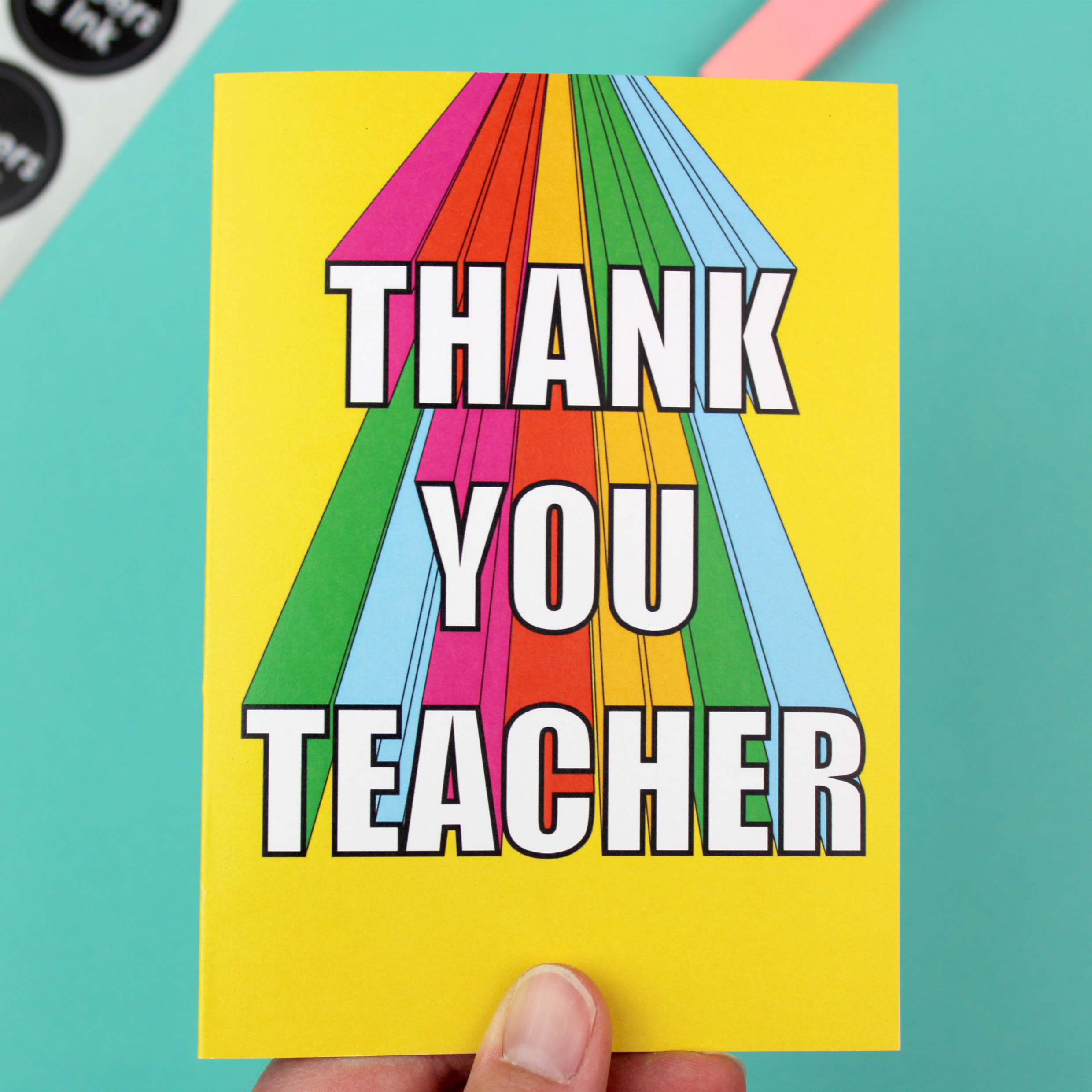 The image shows a bright and colourful Greetings Card and a yellow envelope. The card has a yellow background and rainbow coloured 3D lettering that reads Thank You Teacher.