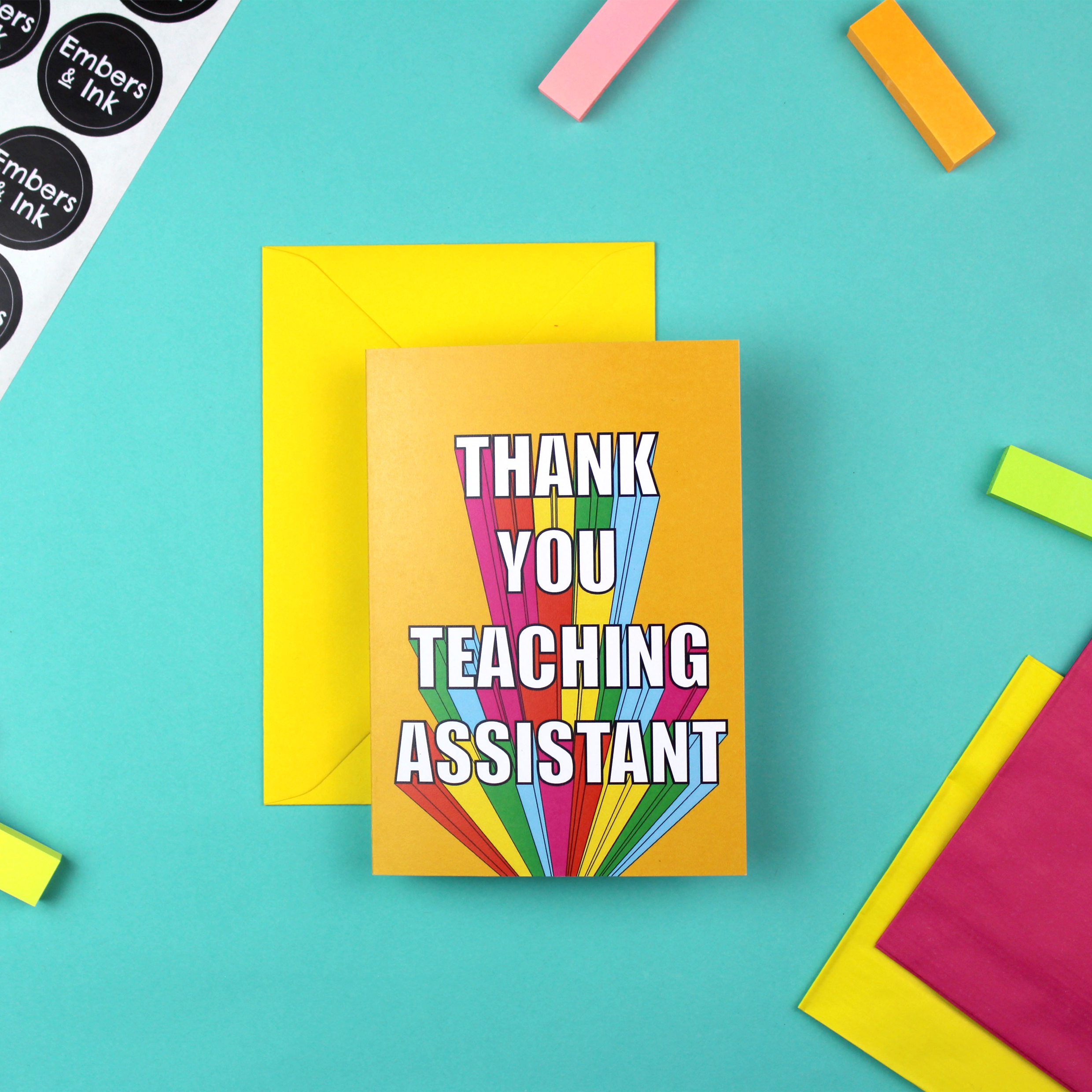 A colourful card with a yellow envelope is shown. The card is orange with rainbow block lettering that reads Thank You Teaching Assistant.