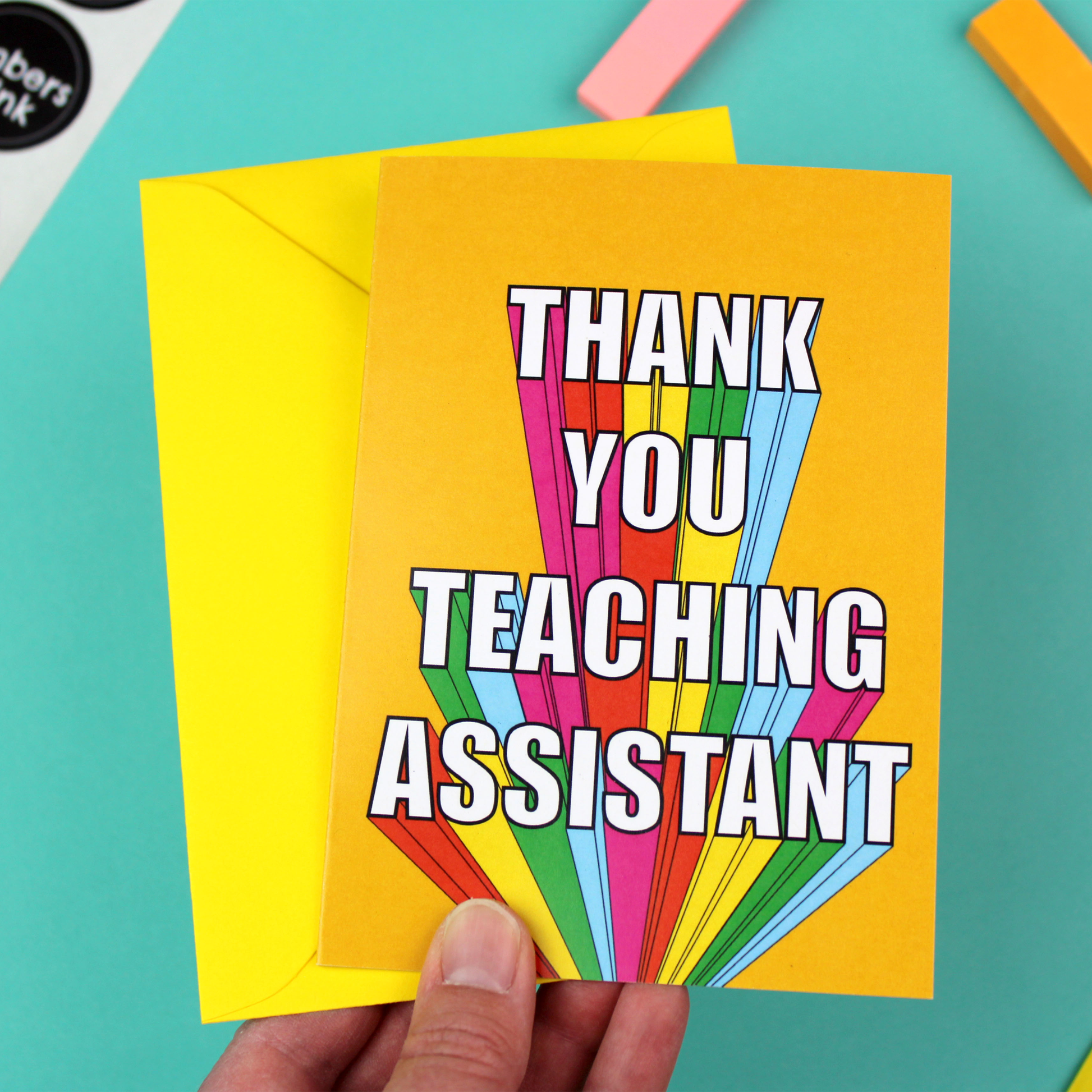 A colourful card with a yellow envelope is shown. The card is orange with rainbow block lettering that reads Thank You Teaching Assistant.