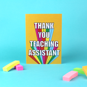 A colourful card is shown standing against a green background. The card is orange with rainbow block lettering that reads Thank You Teaching Assistant.