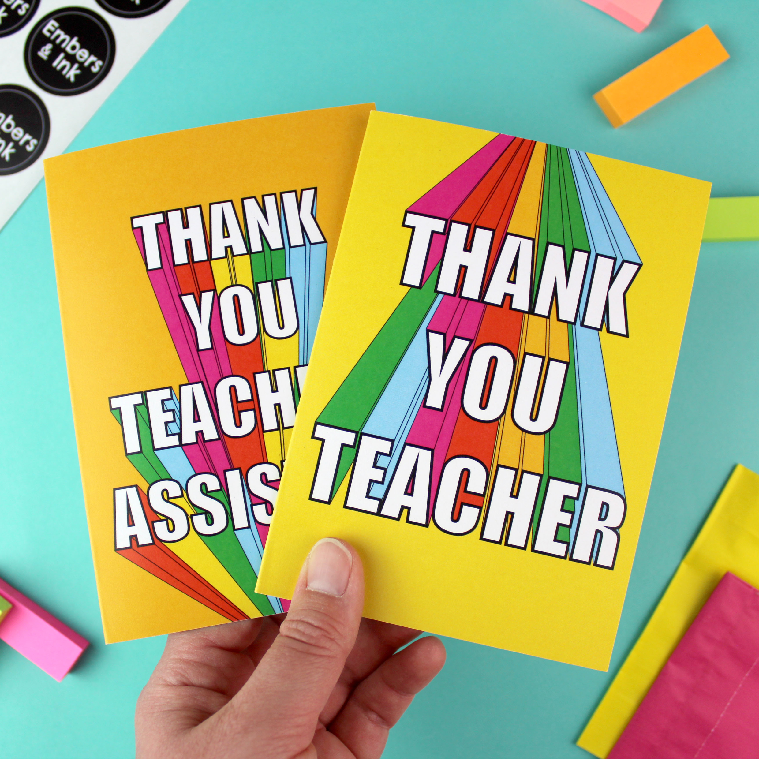 A hand holds two cards, one with a yellow background and one with an orange background. Both cards have rainbow block lettering. One card reads Thank you teaching assistant. The other reads Thank you teacher.