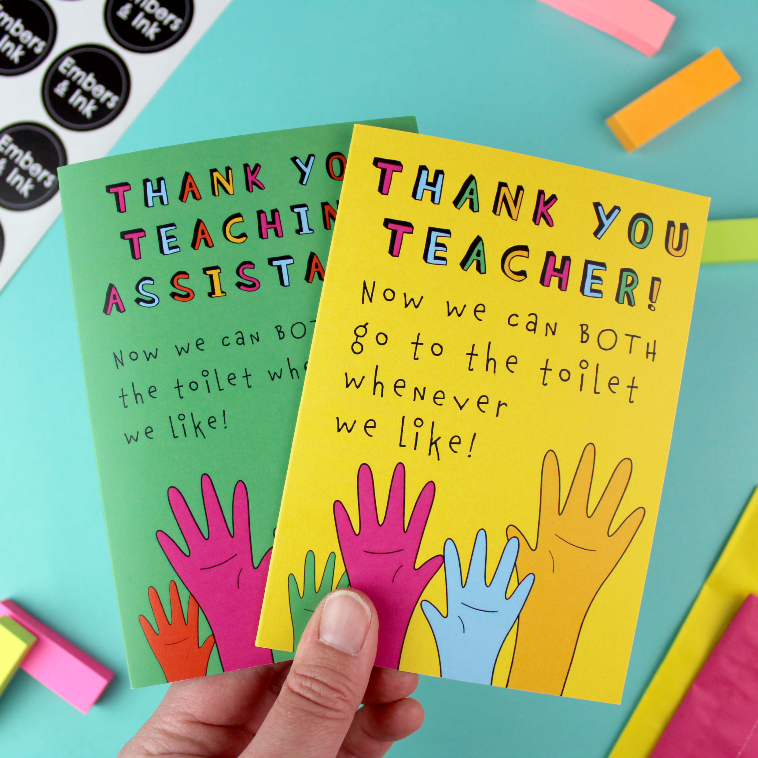 A hand holds two greetings cards, one with a green background and one with a yellow background. Both have an illustration of raised hands of various colours. The yellow card reads Thank You Teacher now we can both go to the toilet whenever we like!. The green card says the same, but for a Teaching Assistant.