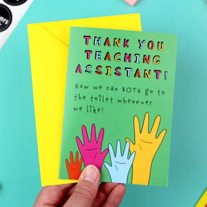 A hand holds a brightly coloured card and a yellow envelope. The card is green an features an illustration of four raised hands in orange pink blue and yellow. Above it are the words 'Thank you teaching assistant! Now we can both go to the toilet whenever we like!'