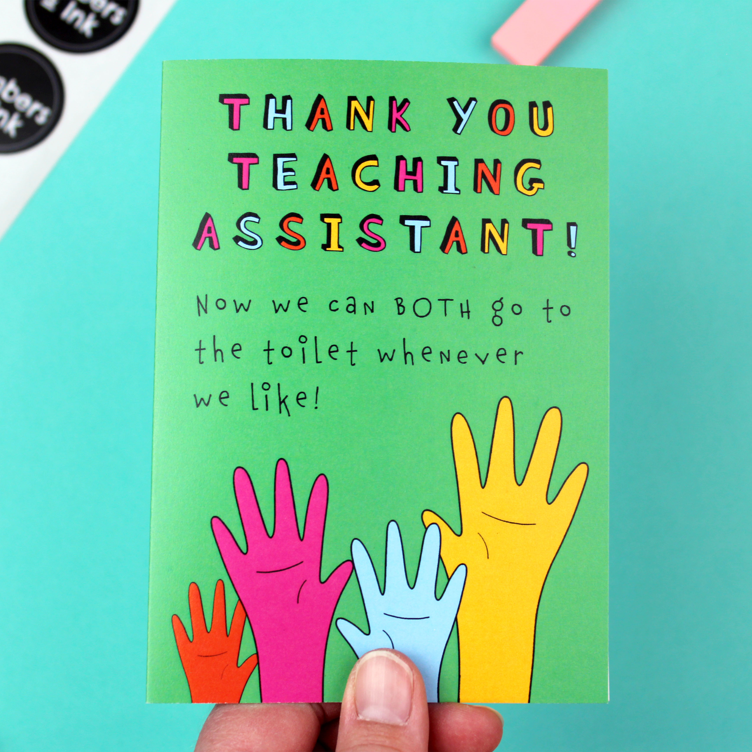 A hand holds a brightly coloured card. The card is green an features an illustration of four raised hands in orange pink blue and yellow. Above it are the words 'Thank you teaching assistant! Now we can both go to the toilet whenever we like!'