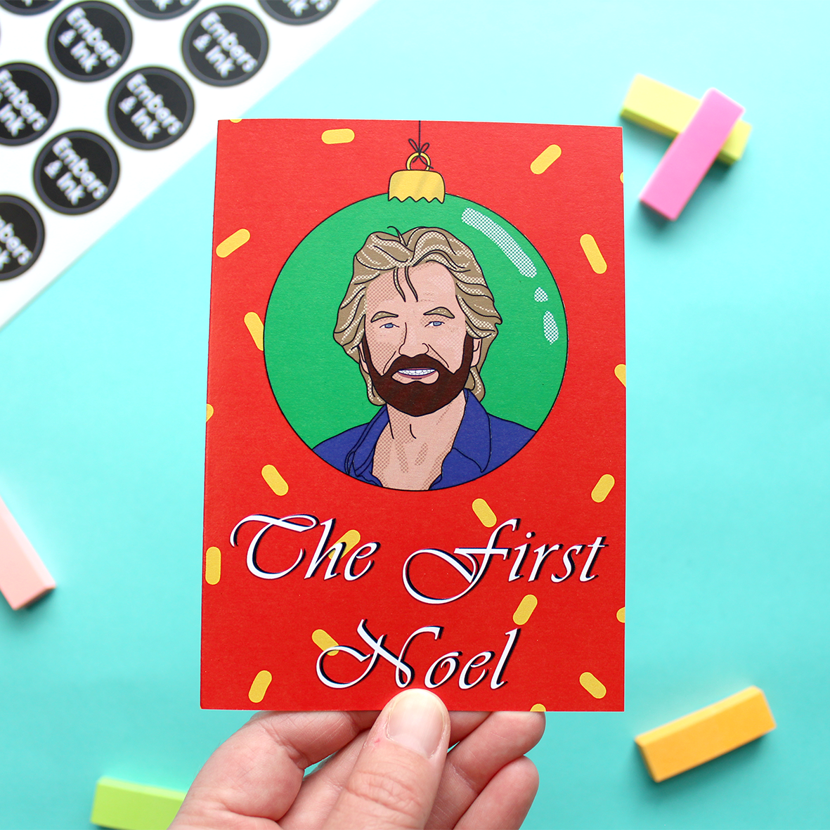 A hand holds a Christmas card showing the face of noel Edmonds in a bauble above the words the first noel. The image is an illustration. The background is red with yellow shapes on it and the bauble has a green background.