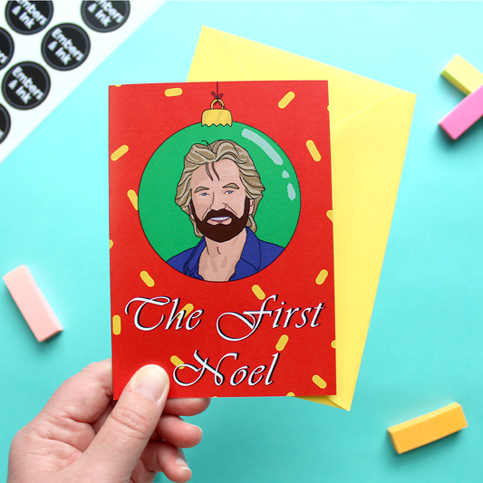 A hand holds a Christmas card showing the face of noel Edmonds in a bauble above the words the first noel. The image is an illustration. The background is red with yellow shapes on it and the bauble has a green background.