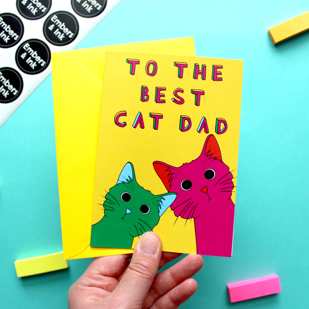 A hand holds a yellow greetings card that has an illustration of two colourful cats (green with blue eard on the left and pink with orange ears on the right. The cats are underneath the colourful words 'To the best cat dad' The hand also holds a hellow envelope and in the background can be seen a sheet of Embers and Ink stickers.