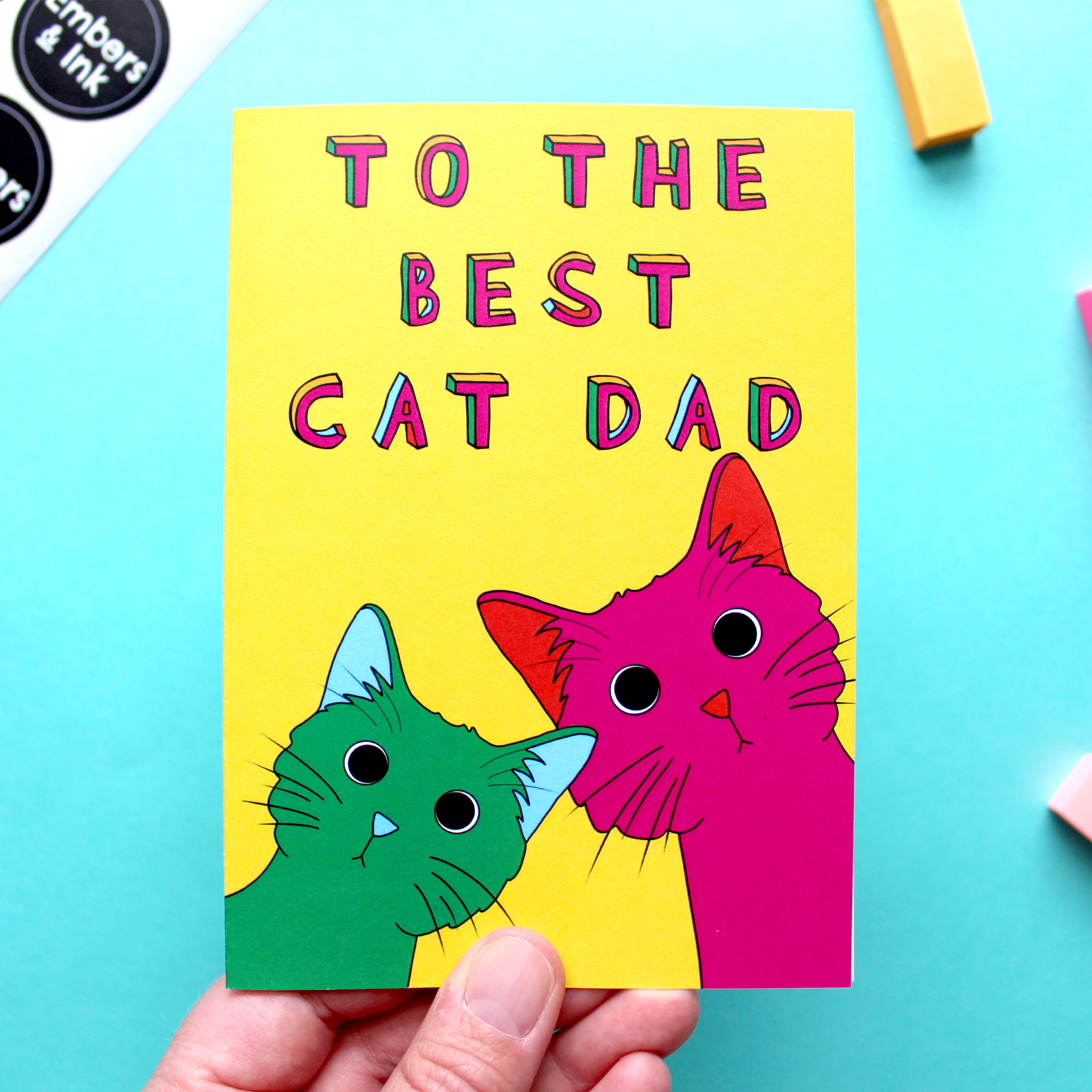 A hand holds a colourful greetings card that has an illustration of two cats under the words 'To the best cat dad' the left cat is green with blue inner ears and nose, and the right cat is pink with orange inner ear and nose. They are looking quizzical.