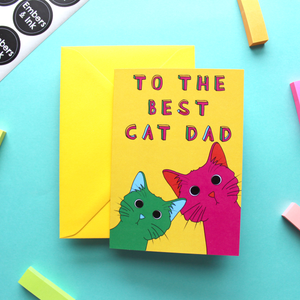 A yellow envelope is shown with a bright greetings card on a colourful background. The cards has a yellow background and an illustration of a green and a pink cat under the words 'To the best cat dad'