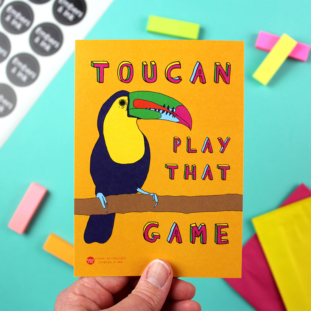 A hand holds a portrait orientated small art print. it has an orange background and an illustration of a toucan bird. next to it are the words 'Toucan plat that game'.