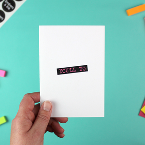 A hand holds a white greetings card with blank and pink text in the style of a dymo label writer. The text reads 'You'll do' in fink font with a black boxed background.