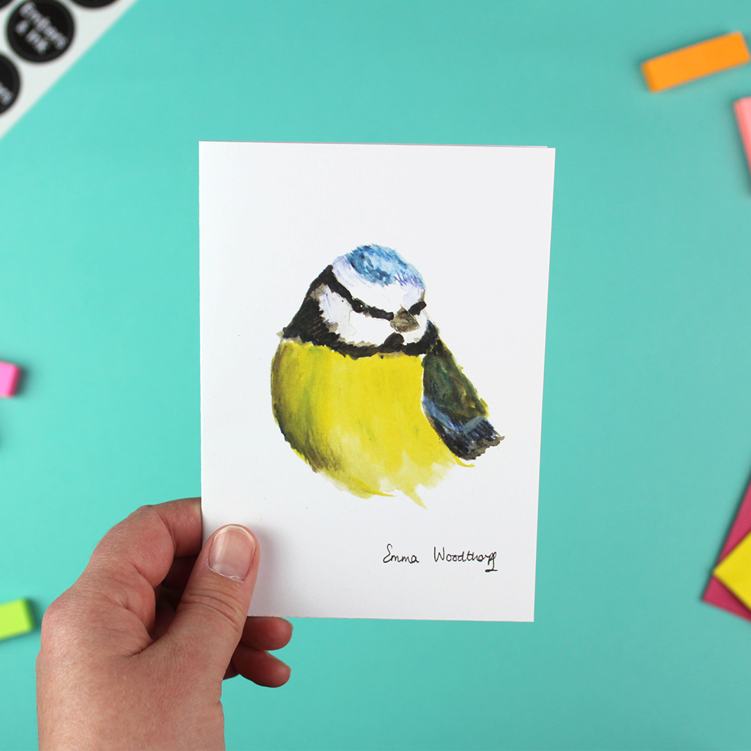 A hand holds a portrait orientated A6 greetings card with a watercolour image of a blue tit bird.