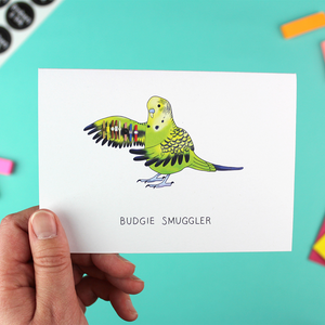 A hand holds up an A6 Landscape orientated greetings card with an illustration of a budgie holding out its wing that has watches attached. Underneath are the words budgie smuggler.