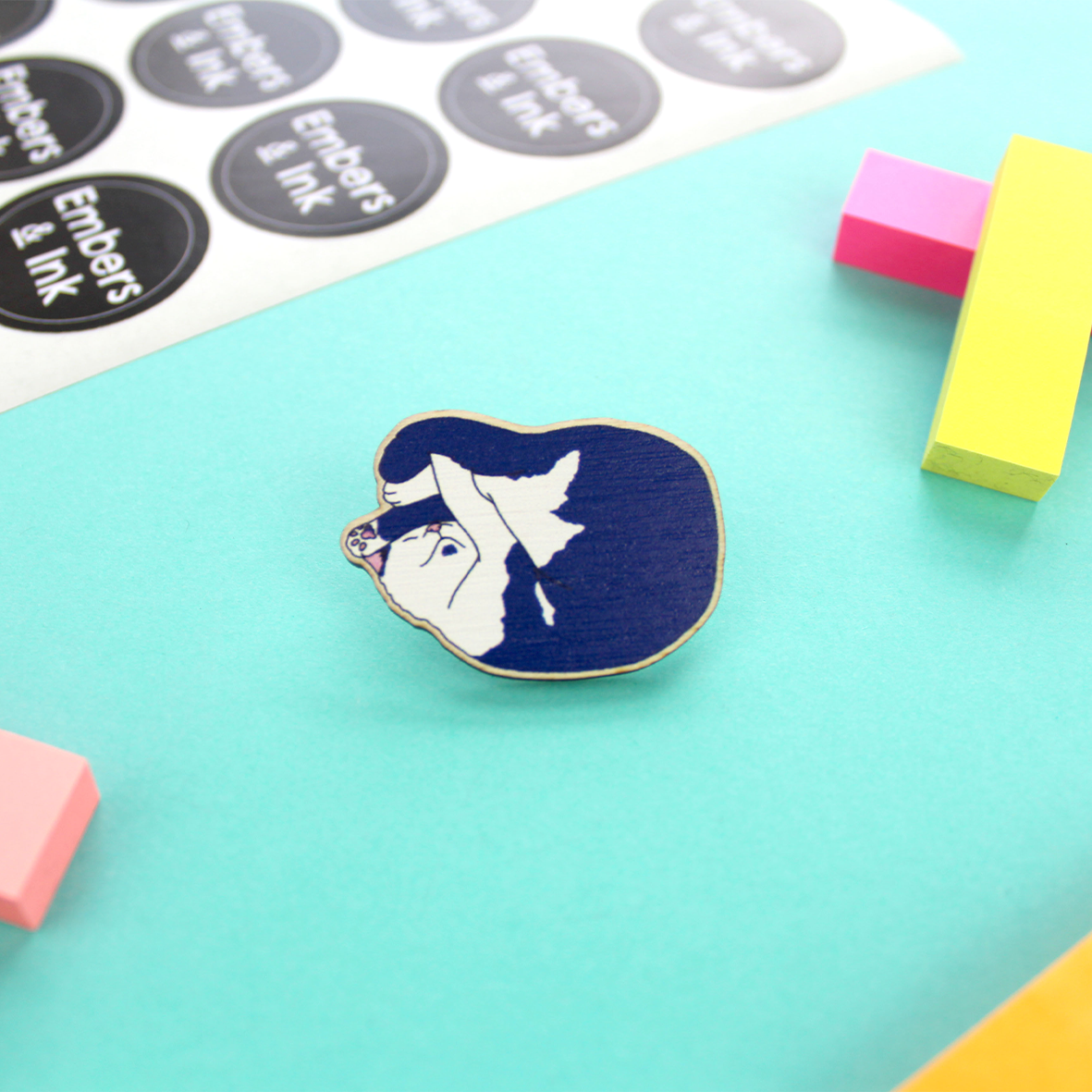 A wooden pin featuring a cute sleeping cat that is all curled up is shown on a table. The cat is blue and white with a pink nose and is drawn in a realistic style..