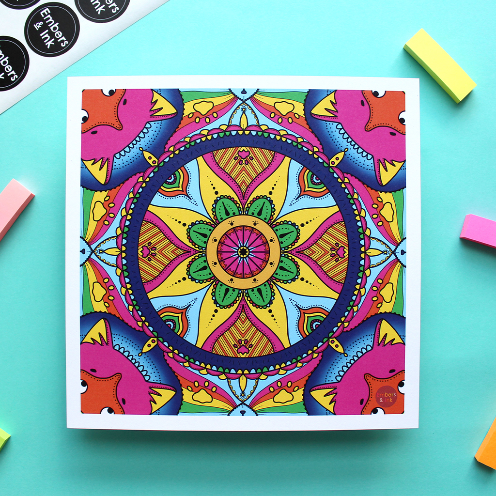 A colourful square art print is shown against a turquise background. The print is a colourful mandala repeating pattern featuring feline things, like cat noses, whiskers, paws, ears and more all hidden in the patternn