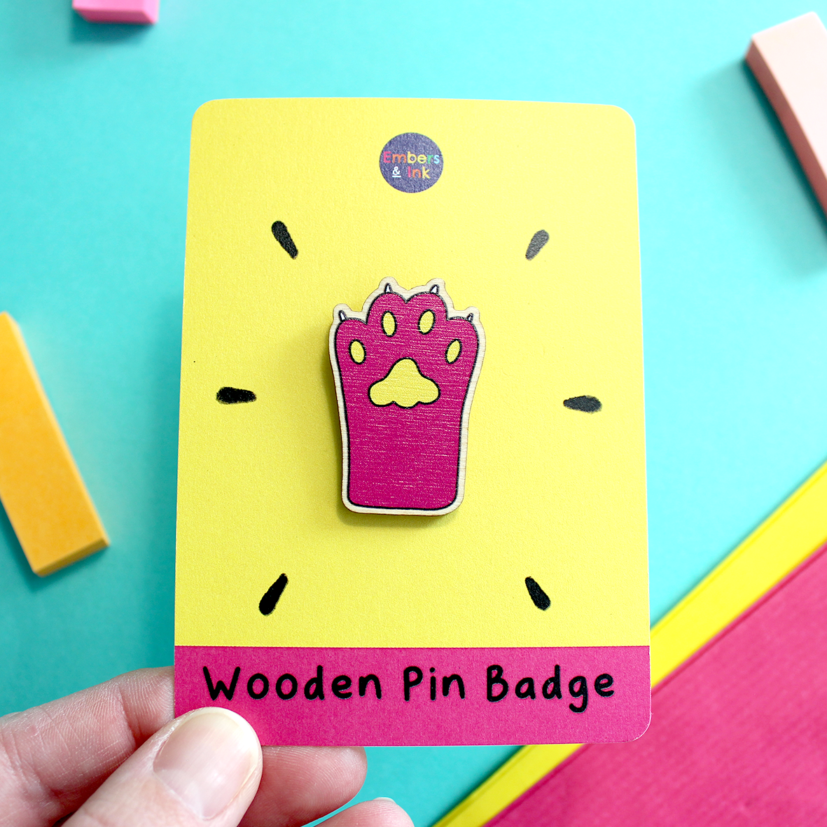 A hand holds a pin paw wooden pin badge that is displayed on a pink and yellow cardboard backer. At. the bottom, on the cardboard backer, are the words Wooden Pin Badge. the Pink cat paw badge has yellow pads and white claws. It is cute!