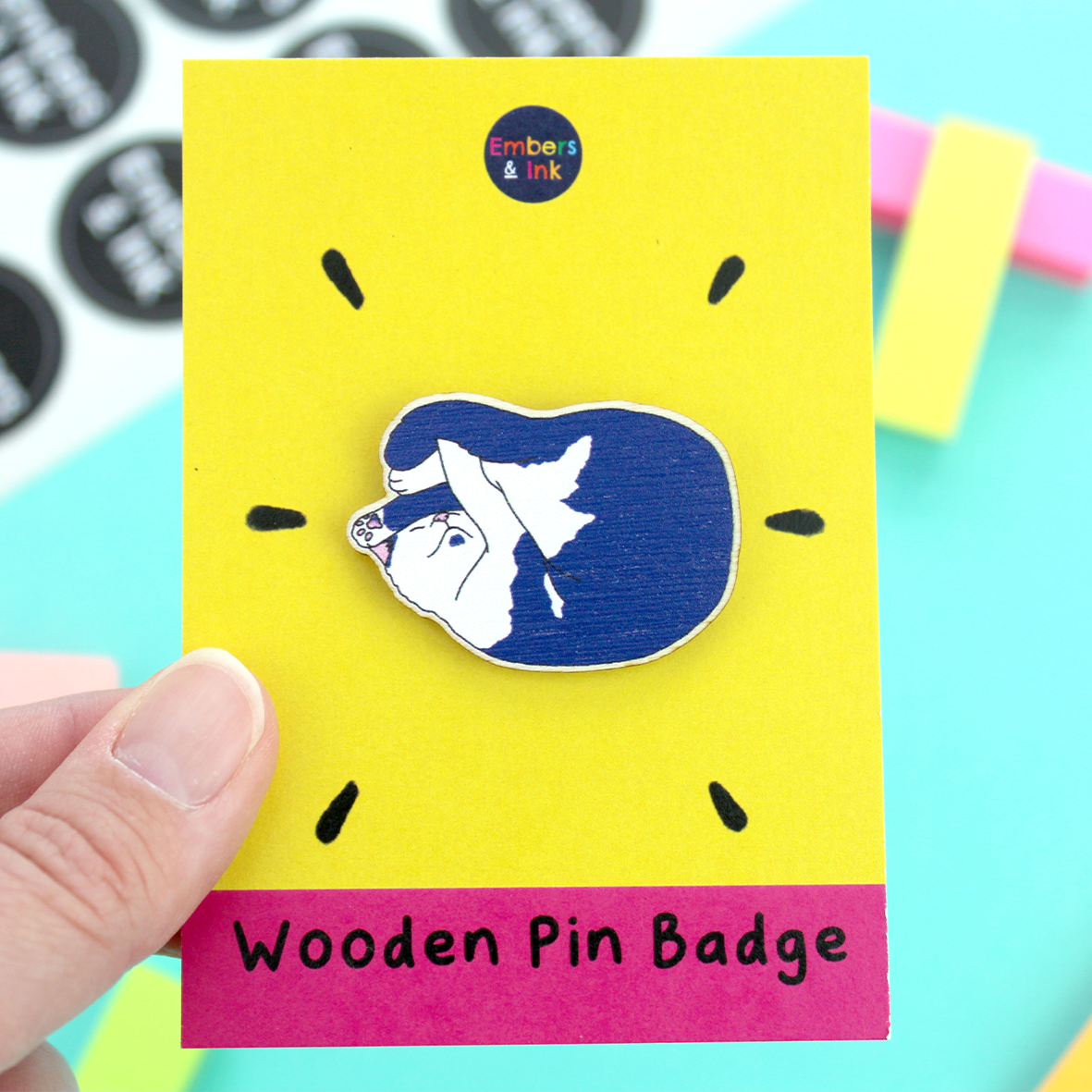 A hand holds the wooden cat pin badge attached to its cardboard backer. The coardboard backer is yellow and pink. the pink section is at the bottom and holds the words 'wooden pin badge'