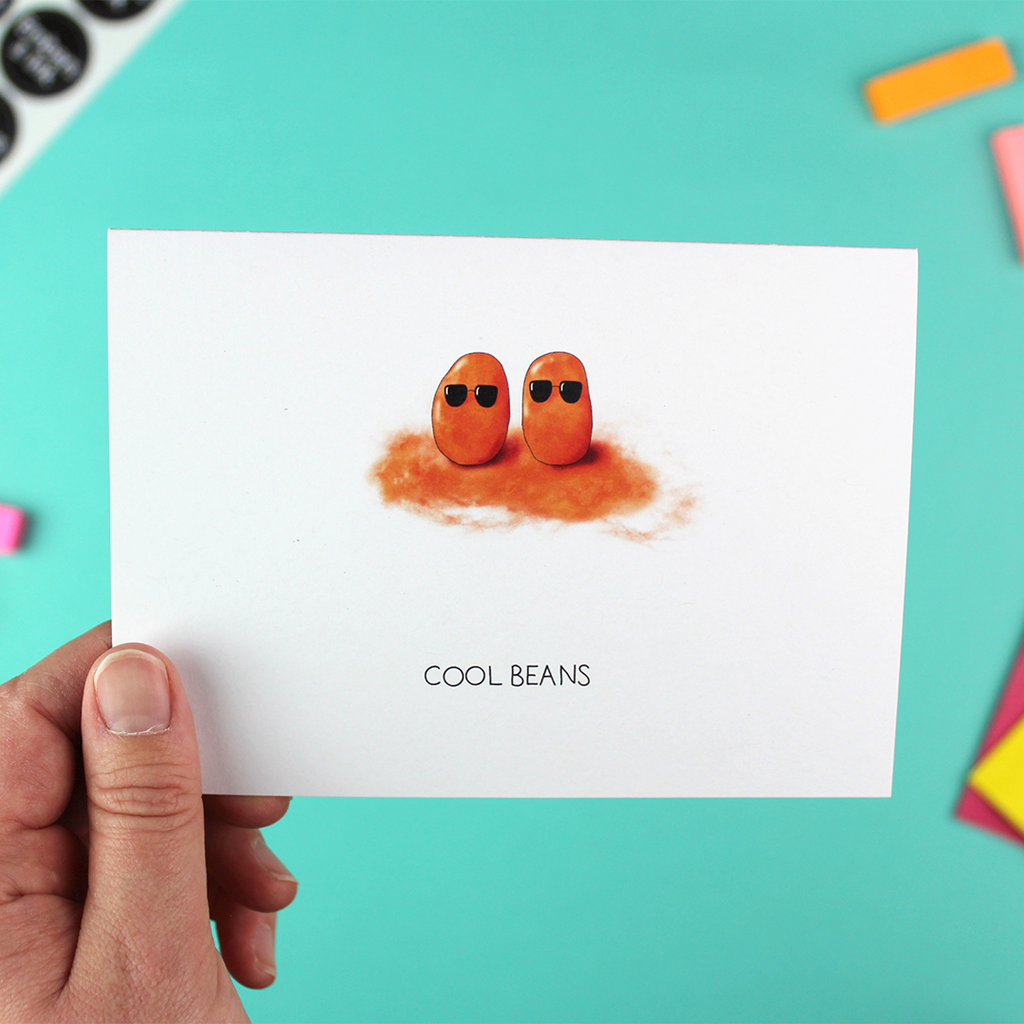 A hand holds a landscape orientated greetings card that shows the illustrated image od two baked-beans wearing shades. Underneath are the words 'cool beans'.