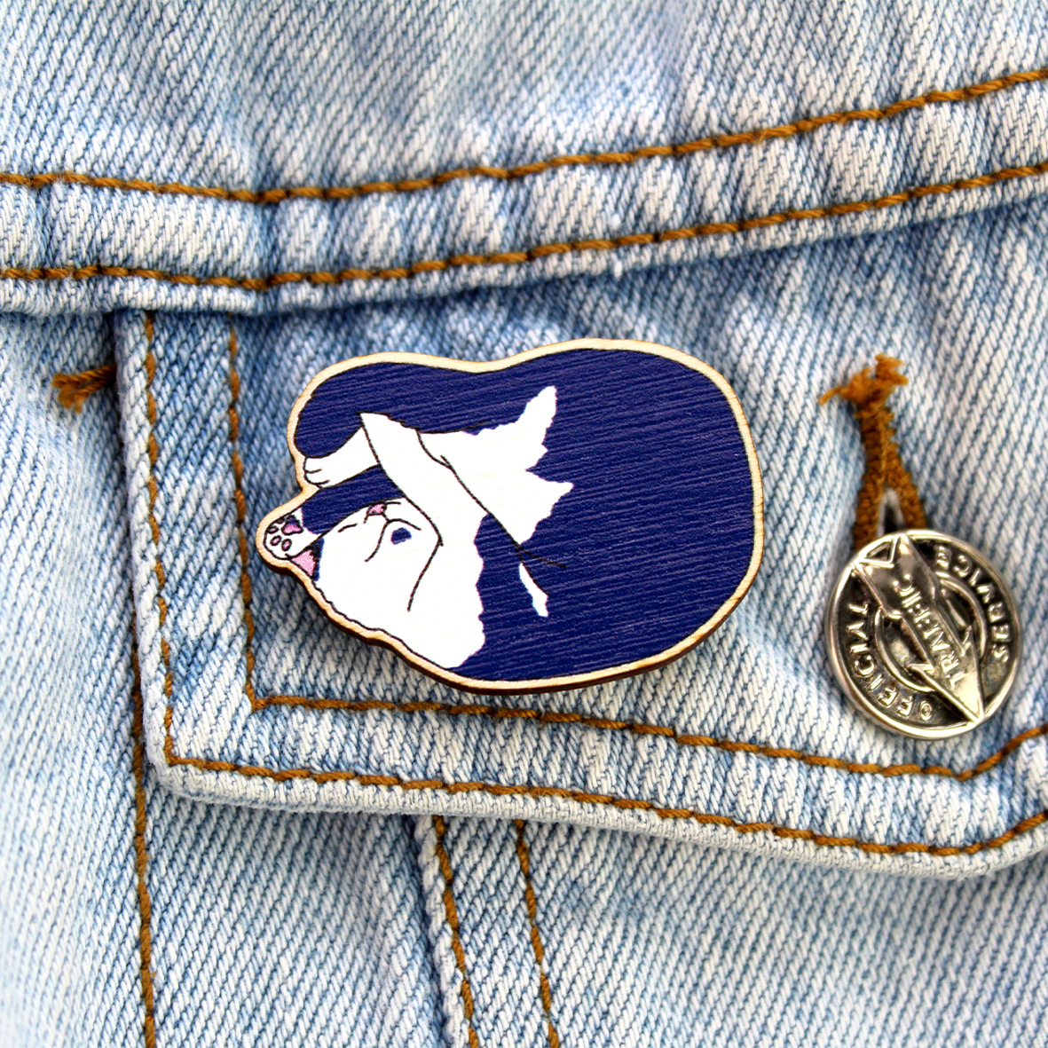 the wooden cat pin basge featuring a sleeping cute cat is pictured attached to a denim jacket pocket.