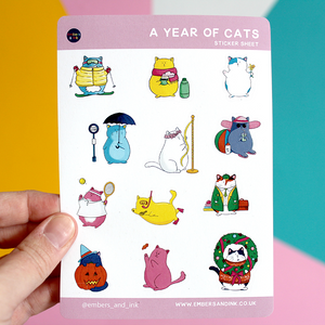 A hand holds up the 'year of Cats' sticker sheet in front of a colourful wall. There are 12 illustrated kiss-cut cats, all doing an activity from a particular month. For example one is putting up a Christmas wreath. One is wearing a witches hat next to a pumpkin.
