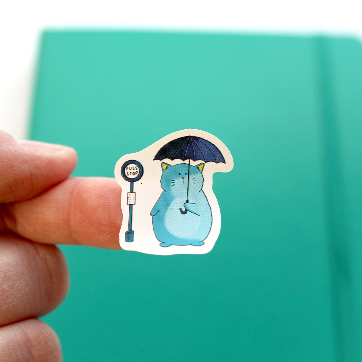 A small blue cat sticker is shown on a finger, It has been kiss-cut. The cat holds an umbrella and has been inspired by Totoro.