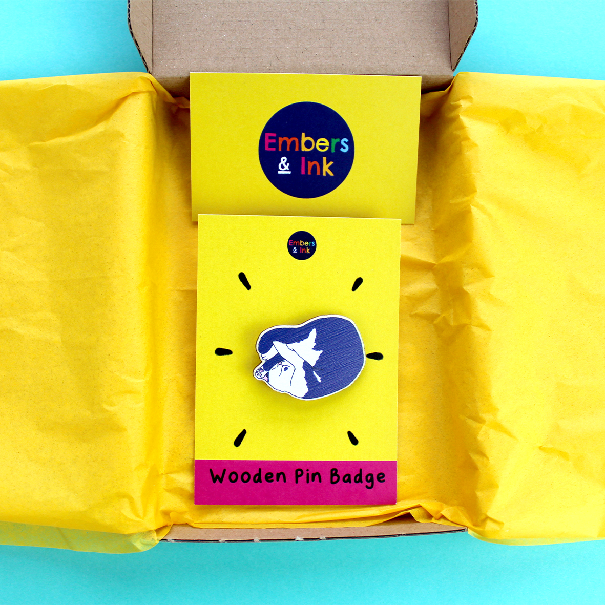 The wooden pink badge is shown attached to its cardboard backer, inside a cardboard box that is lines with yellow tissue paper, with an Embers and Ink business card. It shows that this pin badge is sent plastic-free.
