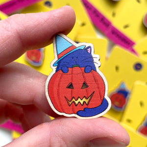 a wooden pin badge featuring an illustrated purple cat wearing a blue witches hat and standing behind a lit spooky halloween pumpkin is shown being held between finger and thumb