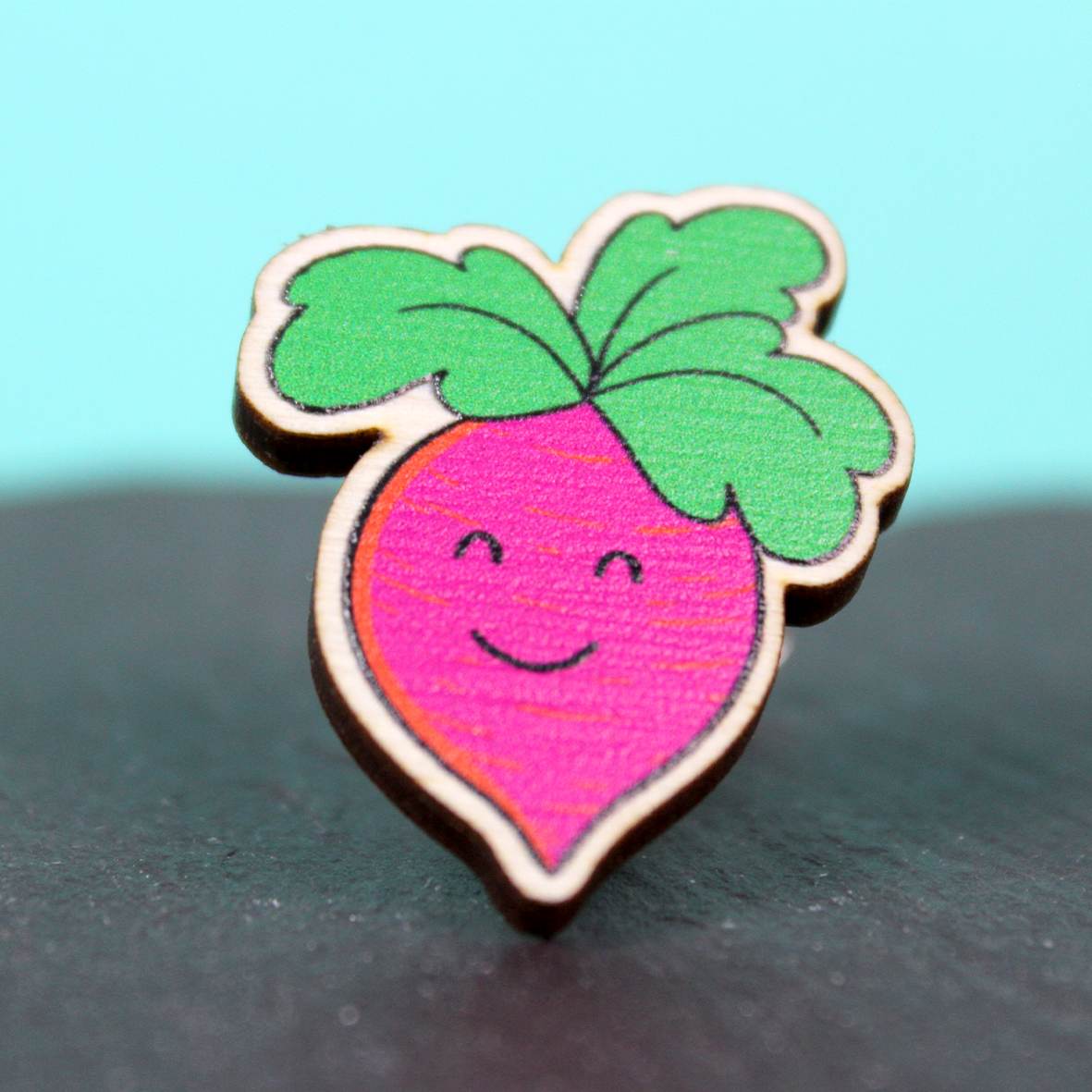 A wooden badge of a pink root vegetable with a smiling face and green leaf hair is shown against a piece of slate