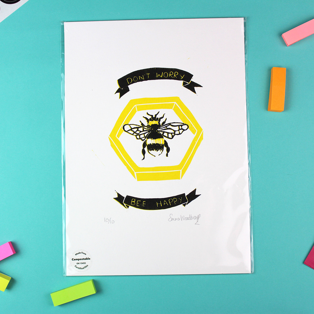 A portrait orientated lino print is shown. it features an image of honeycomb with a yellow and black bumblebee in the middle. above and below are banners containing the words 'don't worry bee happy'