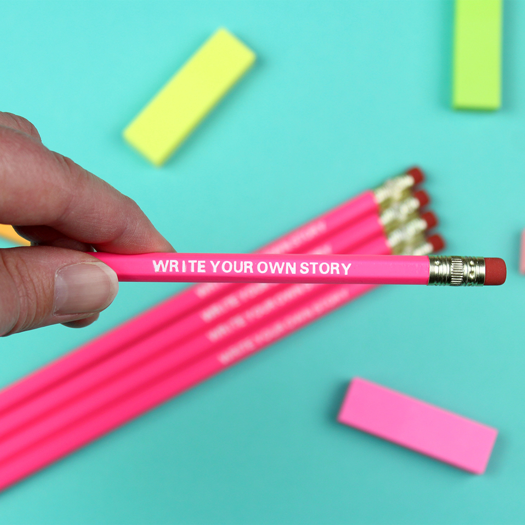A hand holds a bright pink pencil with embossed white lettering reading'write your own story'. 
