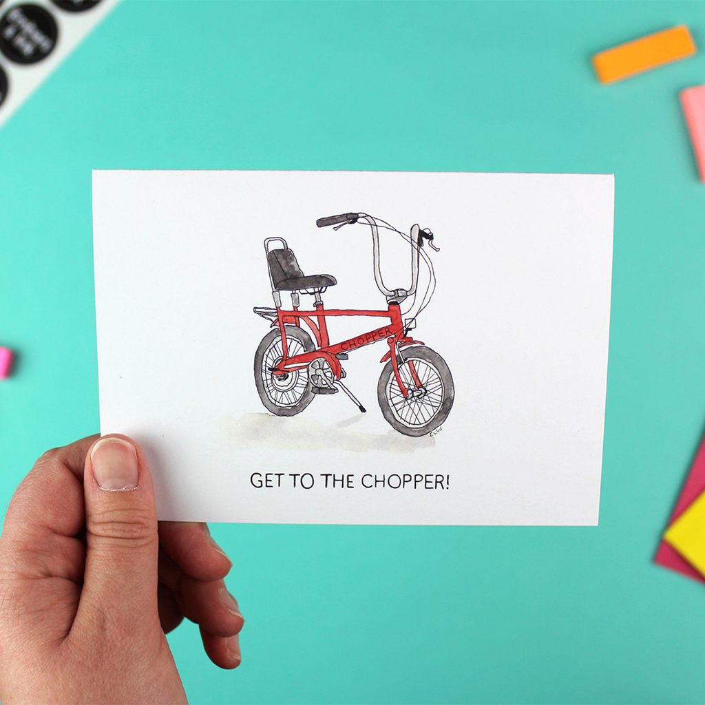 A hand holds a landscape orientated greetings card with an illustration of a red raleigh chopper above the words 'get to the chopper'. the words are also a famous 1980's movie quote.