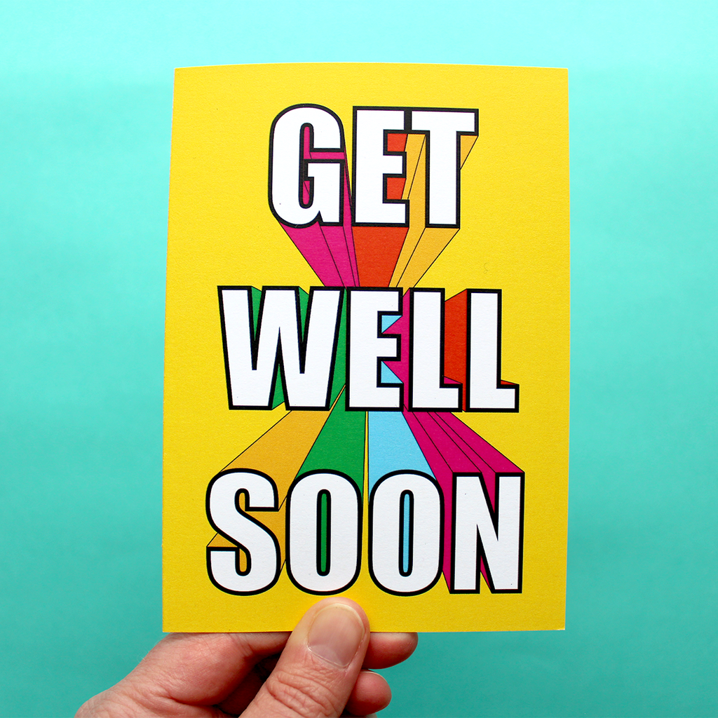 A hand holds a bright and colourful get well soon card. The writing in the middle is bubble writing, and it is also in 3D, with the writing looking like it is flying at you from the middle of the yellow card background. All the sides of the 3D writing is rainbow coloured, making for a very cheerful card for your poorly pal.