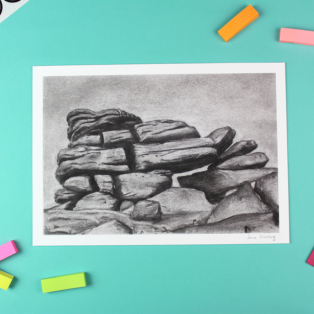 A landscape orientated print of a charcoal drawing lays on a table. The image shows a rock formation.