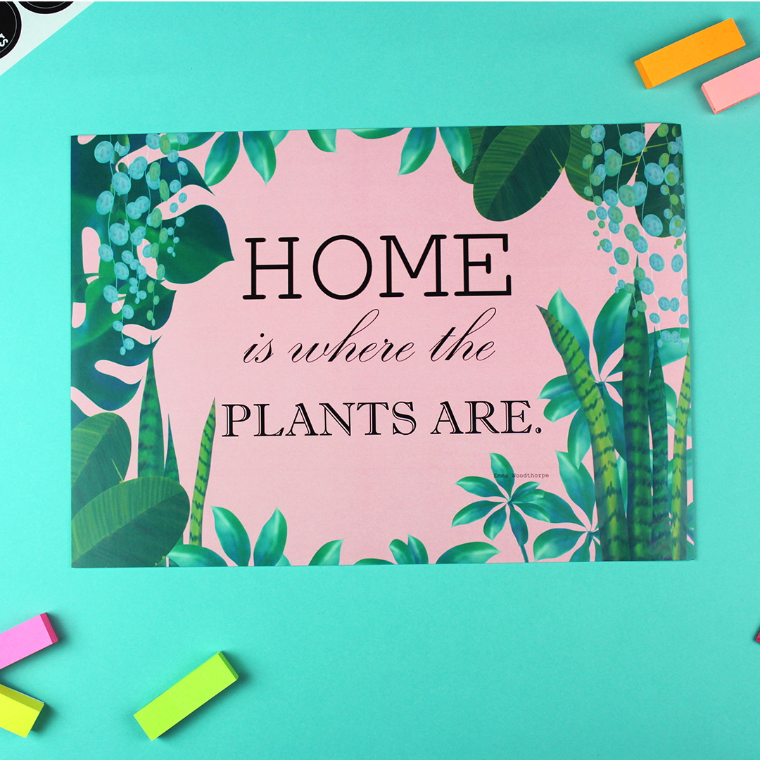 A poster lays on a table. it has a pink background and is framed by green house plants. Inside are the words 'Home is Where the Plants Are'