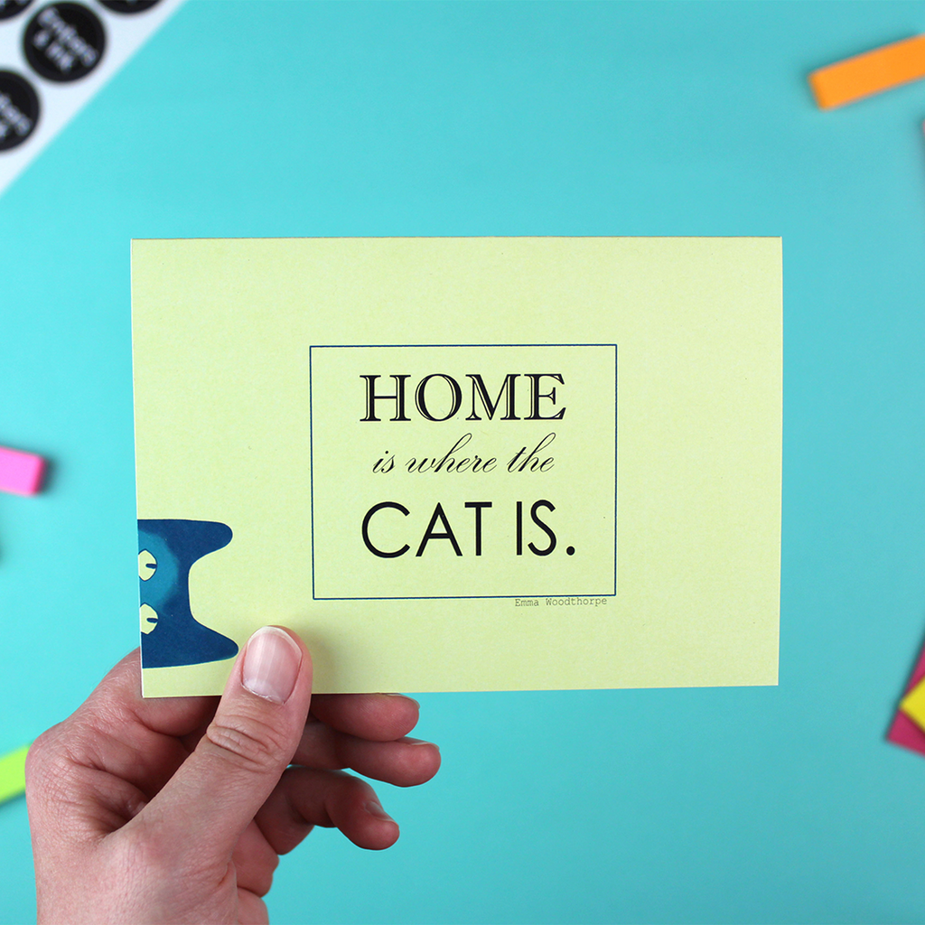 A hand holds up a landscape orientated greetings card, with a yellow background. over the left hand corder a blue and green illustration of a cat peeps over the edge. in the miffle are the words Home is where the cat is