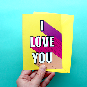 A hand holds a bright and colourful card. The card is yellow with 3D writing in shades of pink and white that say 'I Love You'. The card is held alongside an envelope.