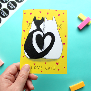 A hand holds a greetings card with an illustration of two cats facing away from us, with their tails entwined to make a heart shape. Below them are the words 'Love Cats'. The background of the card is yellow and the cats are surrounded by many small pink hearts.