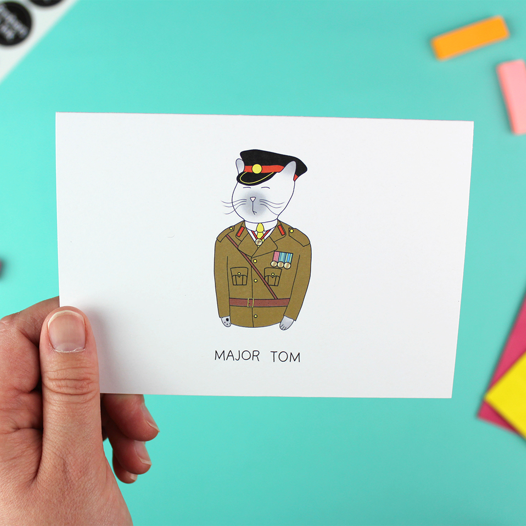 A hand holds up a landscape orientated greetings card with an illustration of a cat in an army uniform. Underneath are the words 'major tom'.