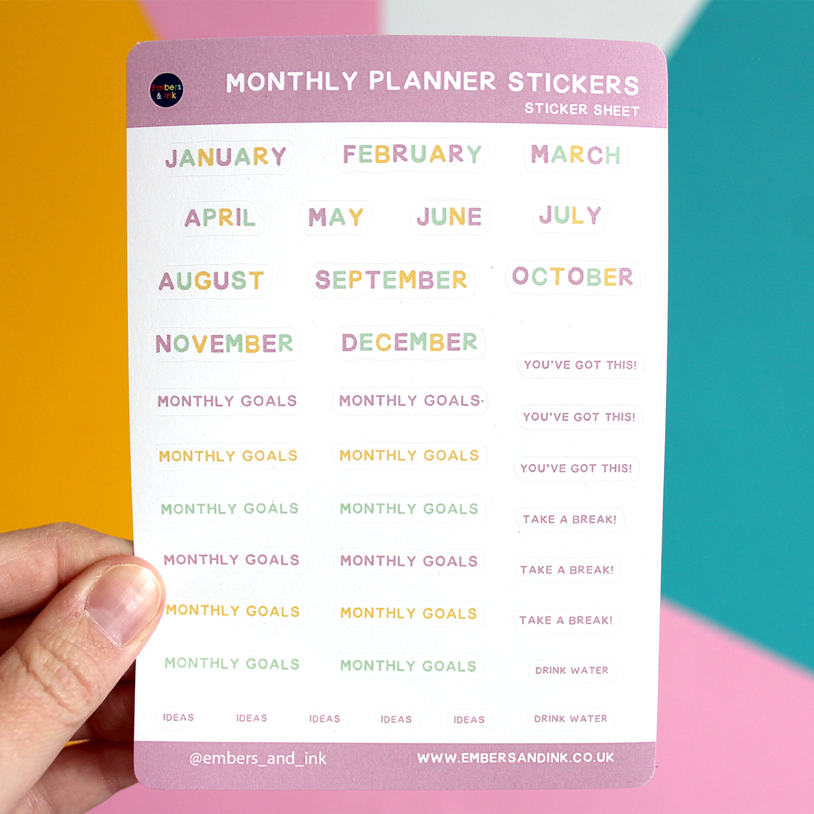 A hand holds the Monthly Planner sticker sheet in front of a colourful wall. The months of the near have their own stickers, plus  12 'monthly goals' stickers. There are also stickers saying 'You've got his' ' take a break' 'drink water' and 'ideas'.