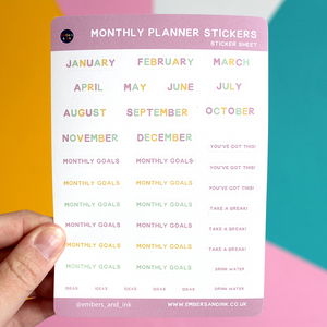 A hand holds the Monthly Planner sticker sheet in front of a colourful wall. The months of the near have their own stickers, plus  12 'monthly goals' stickers. There are also stickers saying 'You've got his' ' take a break' 'drink water' and 'ideas'.